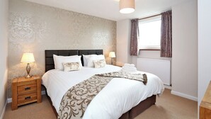 Una cama bien hecha con sábanas blancas y almohadones decorativos, una mesita de noche con lámparas y una manta estampada.