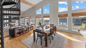 Amplia sala de estar con una mesa de comedor de madera, asientos cómodos, una estantería empotrada y ventanas grandes que ofrecen vistas al exterior.