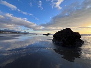 En strand med en stor klippeformation i forgrunden og fjerne bjerge under en delvist skyet himmel.