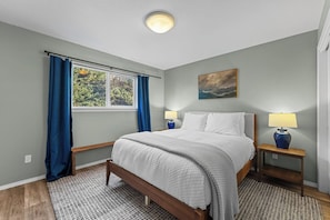 A bedroom with a bed, two bedside tables with lamps, a window with curtains, a painting on the wall, and a ceiling light. - Morning Star Lookout-Spa/Walk to Beach/Ocean Views (Gold Beach)