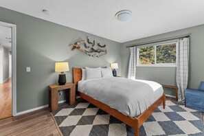 A bedroom with a bed, two bedside tables, a wall-mounted decor piece, a window with curtains, and a view of greenery outside.