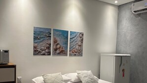 Ein ordentlich bezogenes Bett mit weißer Bettwäsche und einer türkisblauen Decke, eine wandmontierte Klimaanlage und zwei Wandbilder mit Muscheln und Seesternen.