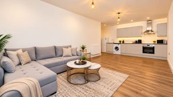 Una sala de estar moderna con un sofá gris, una mesa de centro redonda y una zona de cocina con armarios y electrodomésticos blancos.