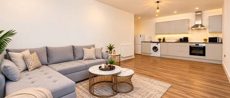 A modern living room with a grey sofa, a round coffee table, and a kitchen area with white cabinets and appliances.