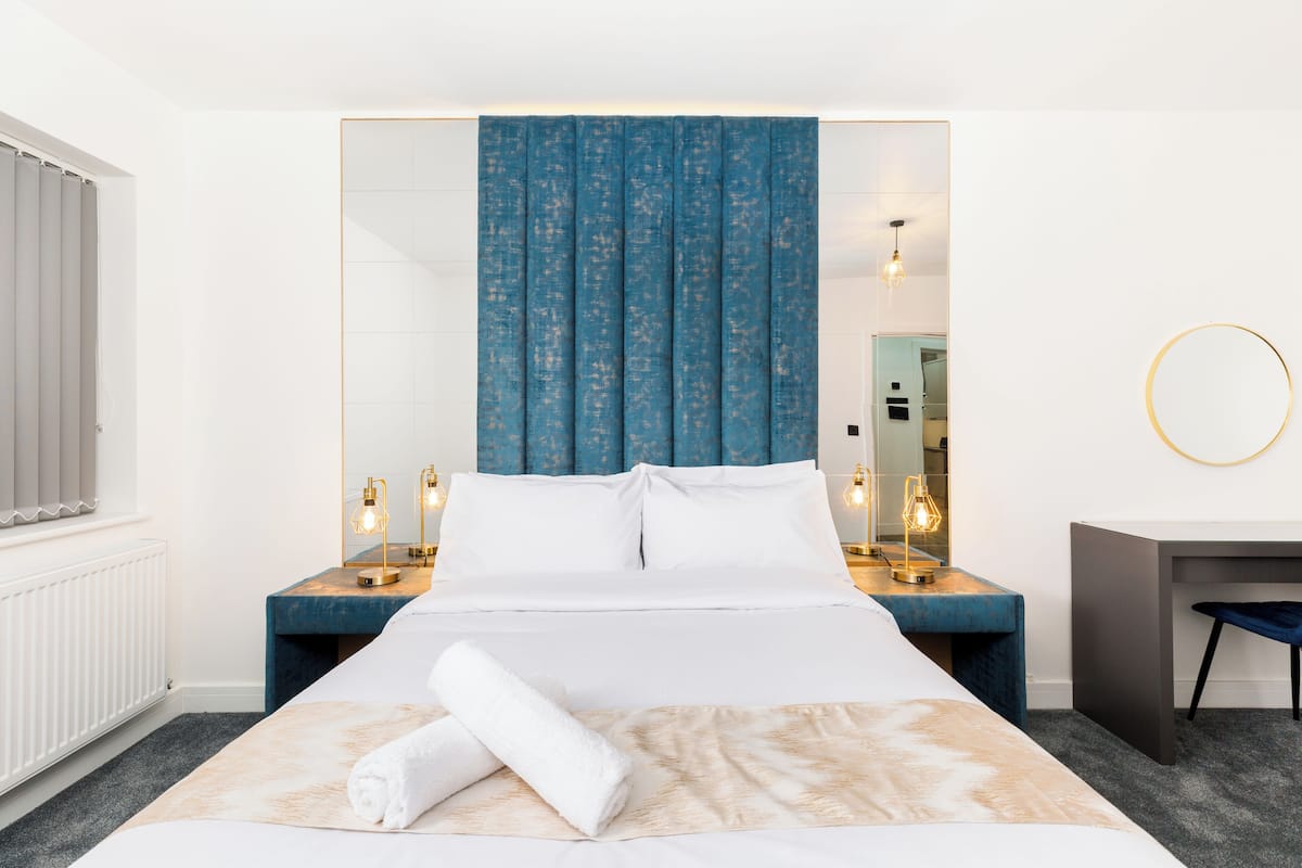 Une chambre d’hôtel avec un grand lit, une table de chevet avec une lampe, un bureau avec une chaise, un miroir rond et une fenêtre avec des stores.