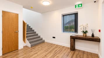 Una escalera conduce a un rellano con una puerta de madera, una ventana con persianas y una mesita consola con una planta y palitos de incienso.