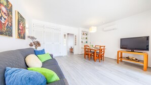 A modern living room with a grey sofa, green and blue cushions, a dining area with wooden chairs and a table, and a flat-screen TV on a wooden stand. - Apartment Portofino 1 (Loulé)