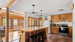 A modern kitchen with wooden cabinets, a stainless steel refrigerator, a microwave, and a stove. There are three black barstools at the kitchen island.