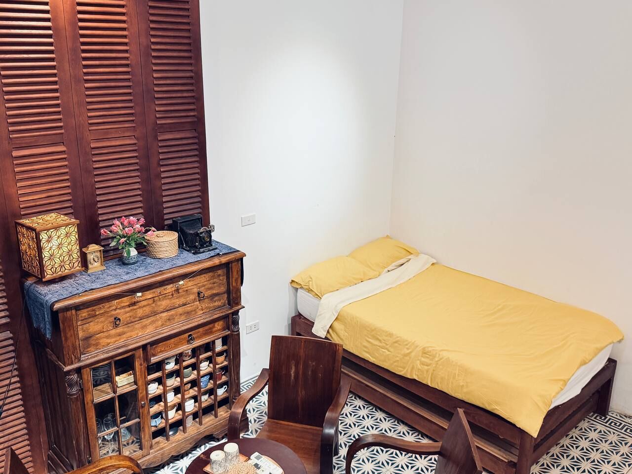 A small, compact room with a single bed, a wooden cabinet with drawers, and a patterned floor.