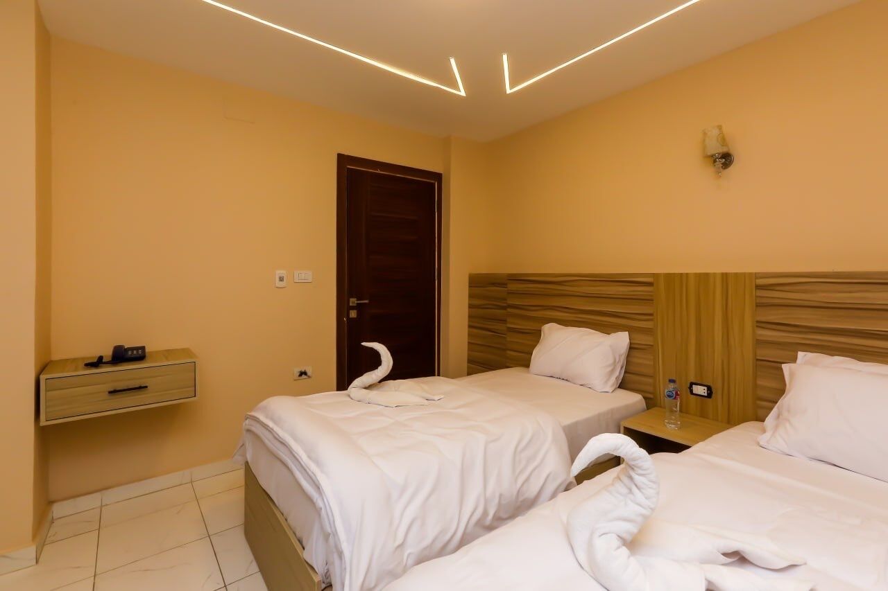 A hotel room with two beds, a wooden headboard, a nightstand with a water bottle, and a wall-mounted light fixture.