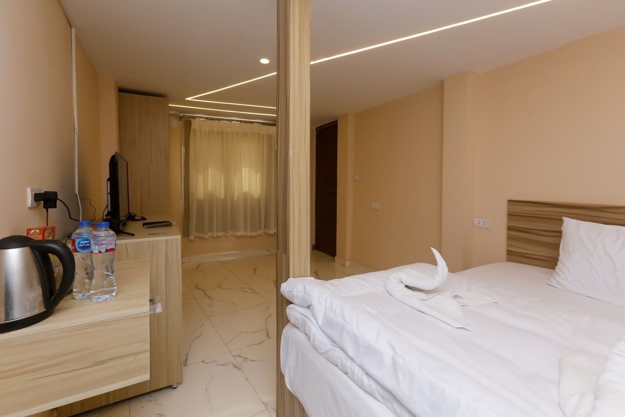 A hotel room with a bed, a wooden desk with a kettle and water bottles, a mirror, and a view of the bathroom.