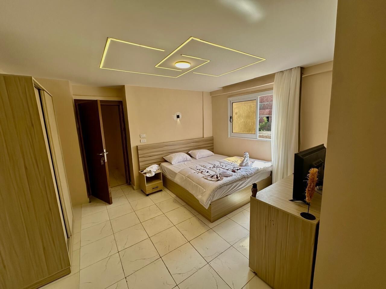 Un dormitorio moderno con cama, mesitas de noche, televisor y ventana con cortinas.