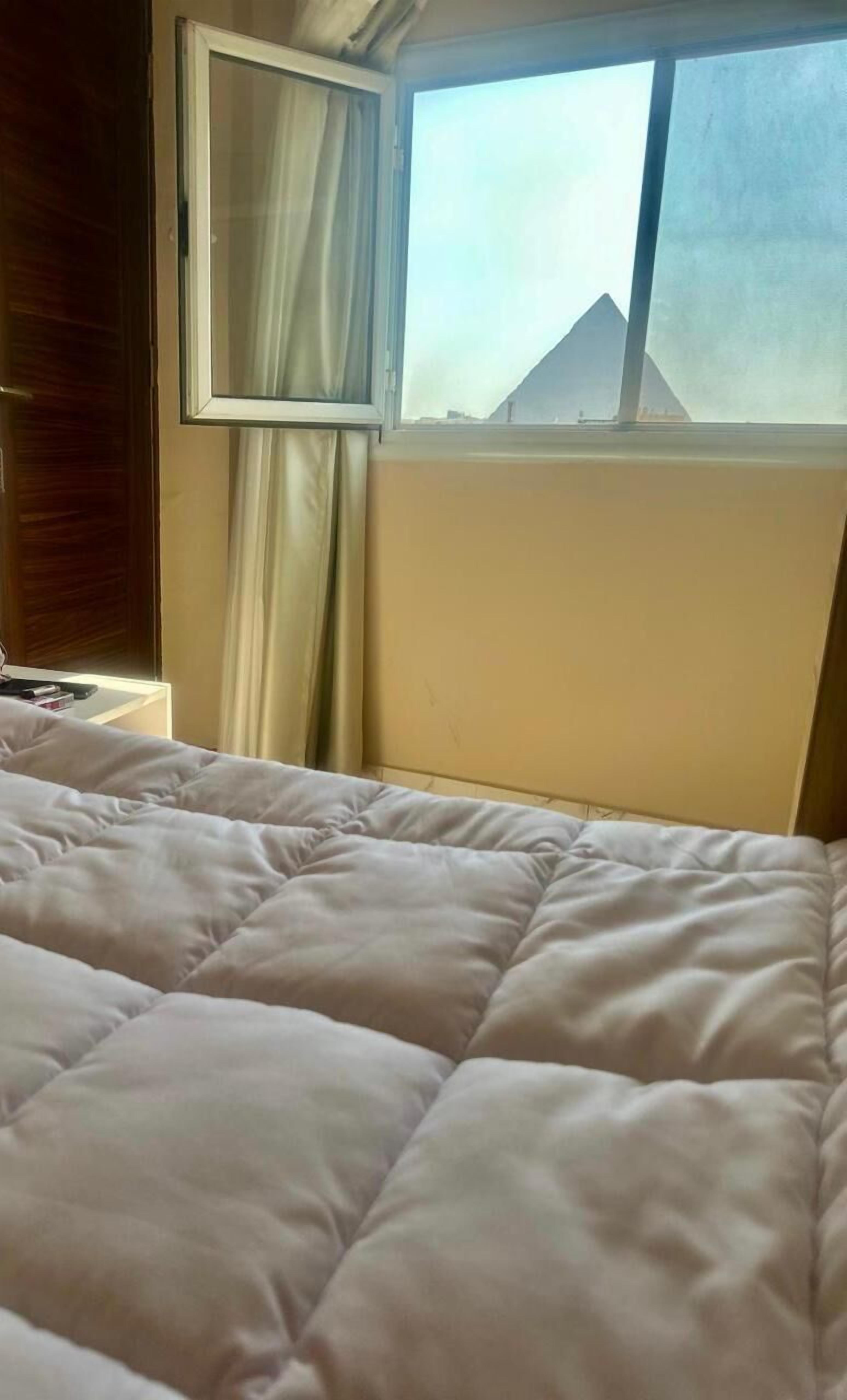 Un lit recouvert d’une couette blanche, une fenêtre avec des rideaux et une vue sur une pyramide.