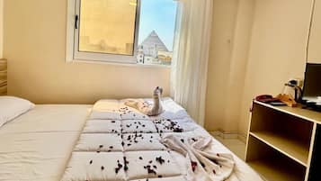 Quarto de hotel com uma cama grande, uma escrivaninha com um computador e uma janela com vista para uma pirâmide.
