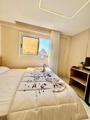 A hotel room with a large bed, a desk with a computer, and a window with a view of a pyramid.