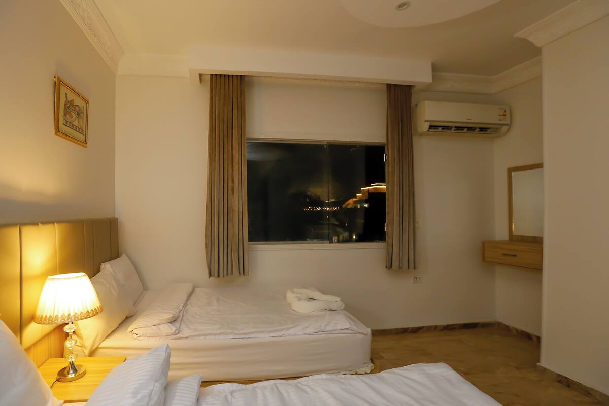 A hotel room with two beds, a nightstand with a lamp, a window with curtains, an air conditioning unit, and a mirror.