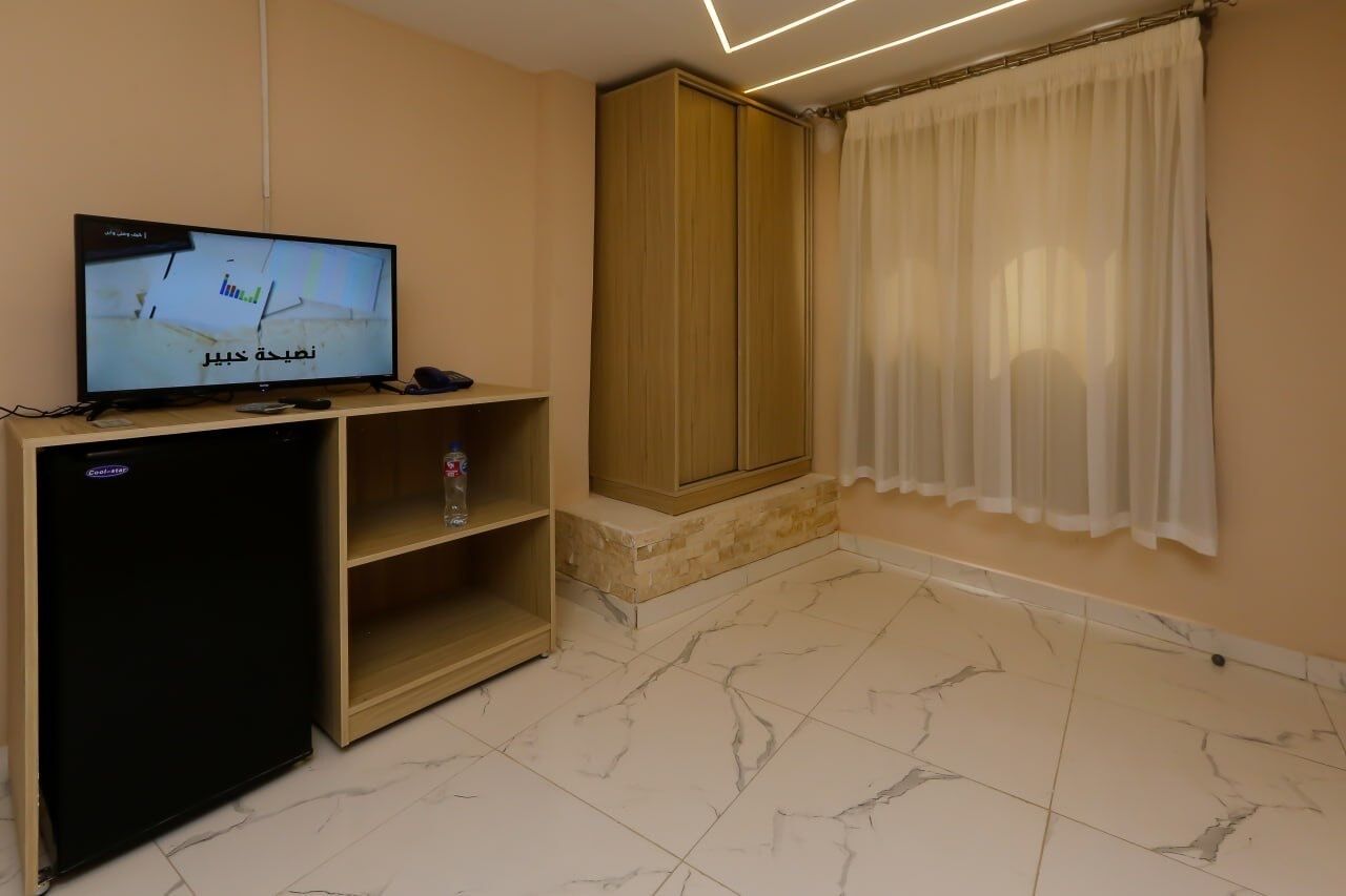 Una habitación con una televisión sobre un soporte, un armario, una estantería con una botella y una ventana con cortinas.