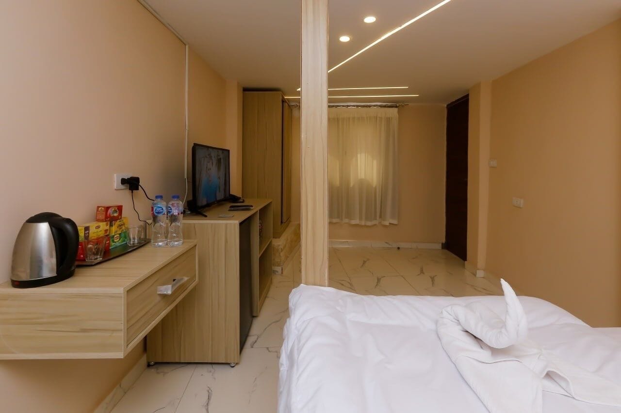 A hotel room with a bed, a desk with a kettle, a TV, and a minibar.