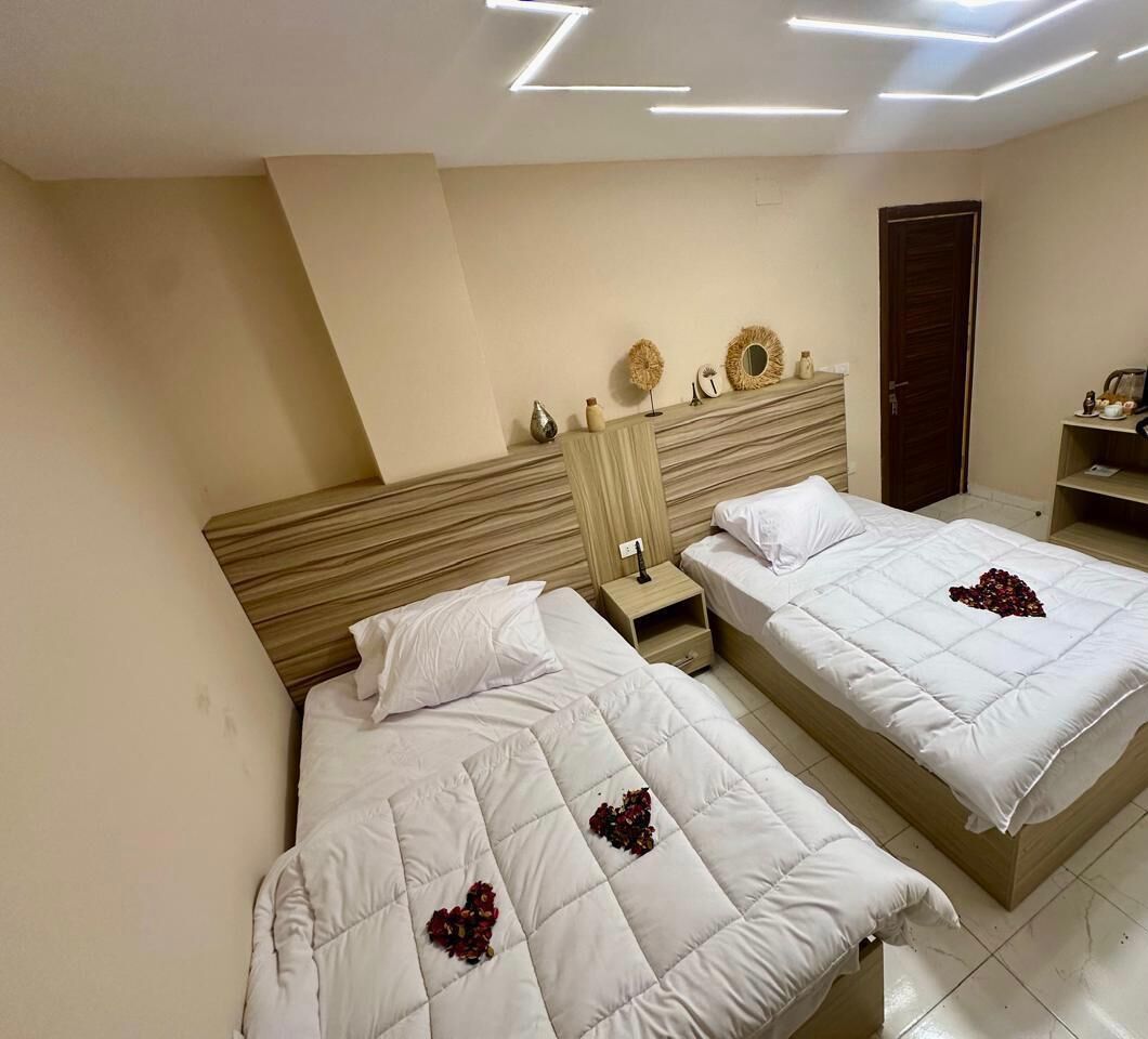 A hotel room with two beds, a wooden headboard, a nightstand, and a wall-mounted shelf with decorative items.