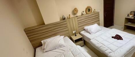 A hotel room with two beds, a wooden headboard, a nightstand, and a wall-mounted shelf with decorative items.