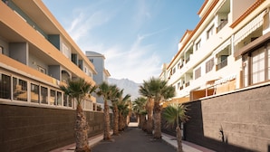 A corridor lined with palm trees and buildings with balconies, leading towards a mountain.