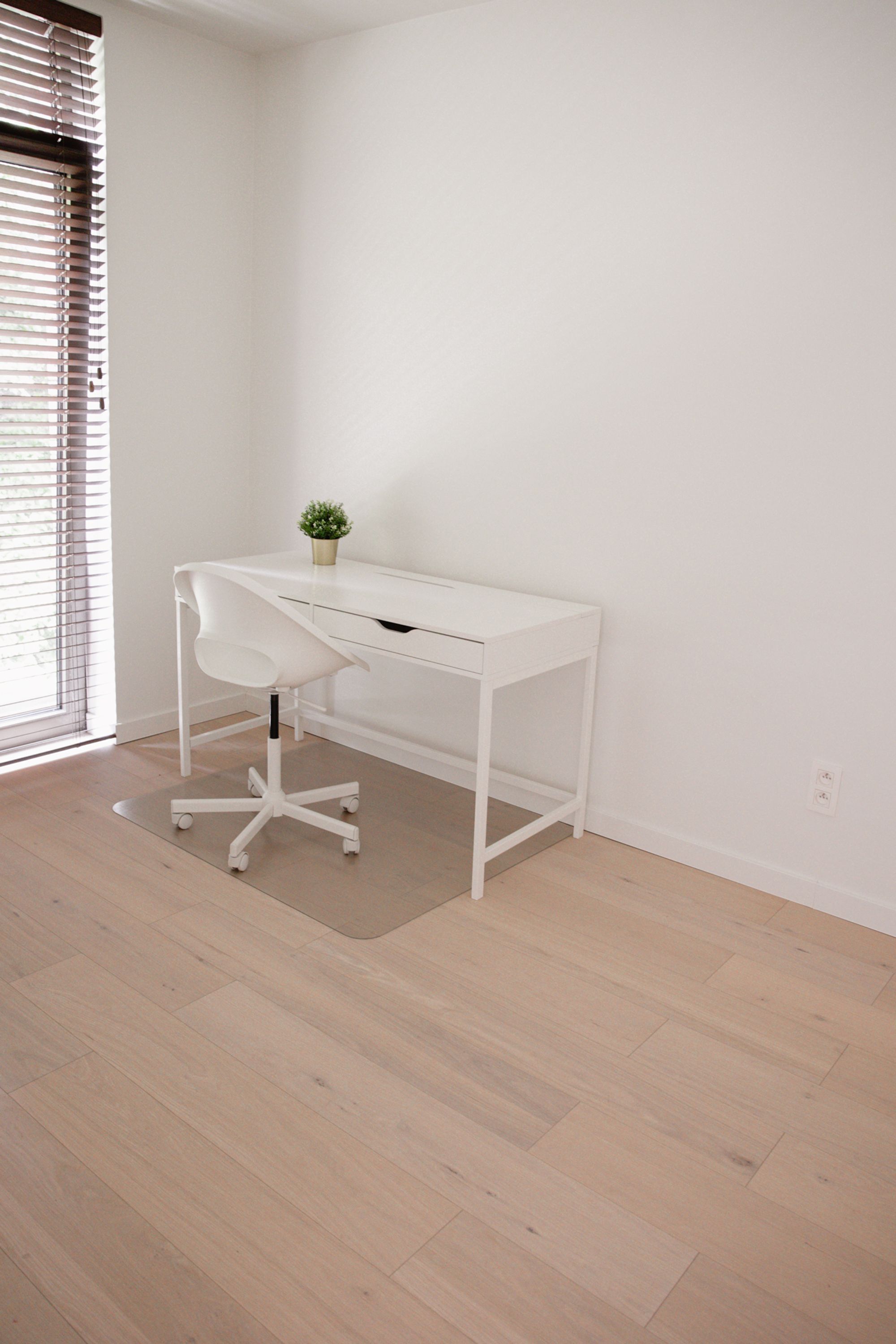 Un bureau blanc orné d’une plante, placé dans un coin d’une pièce dont le sol est en bois et qui possède une fenêtre avec des stores.
