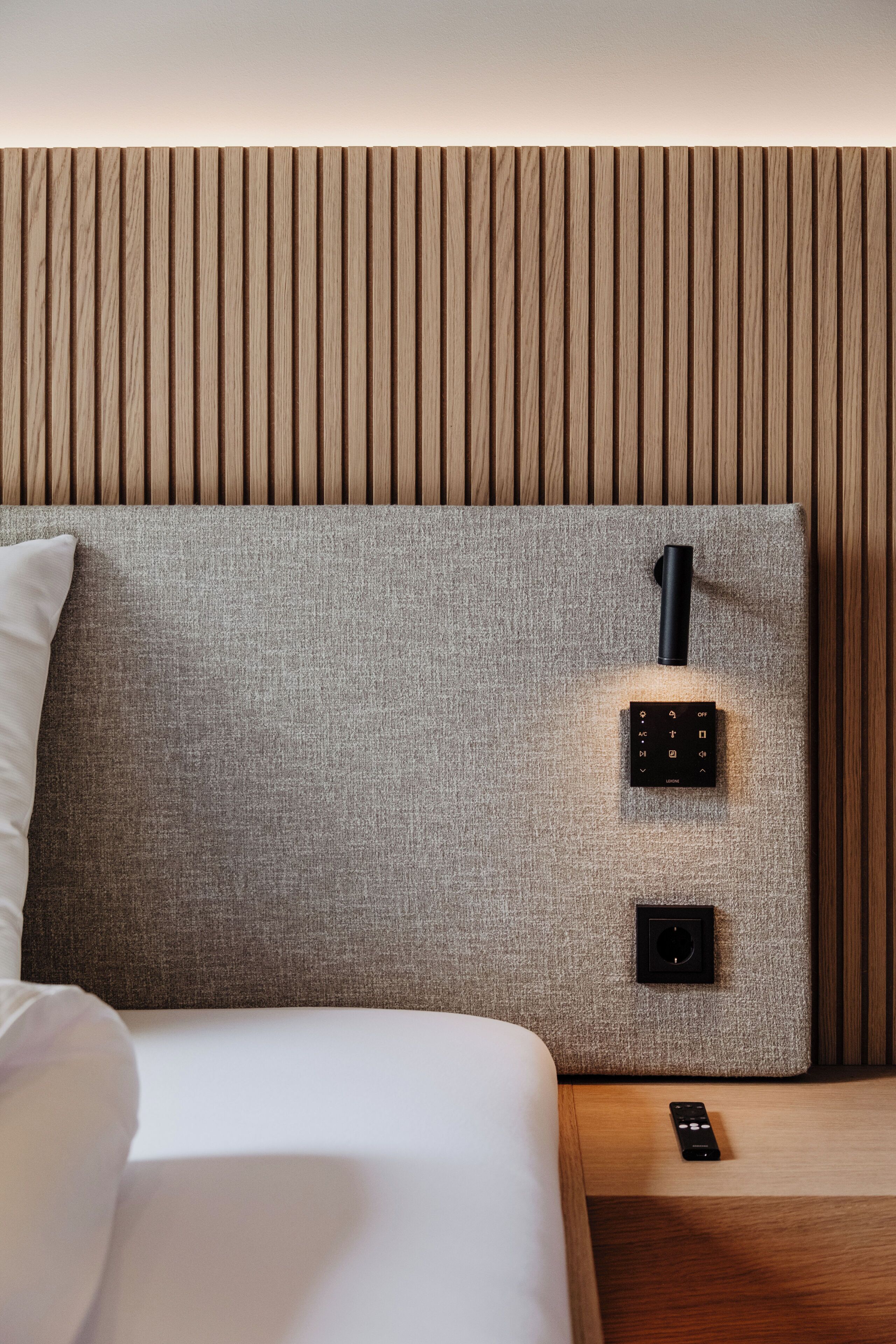 Una cama bien hecha con un cabecero gris, una mesita de noche con un control remoto y una lámpara de pared.