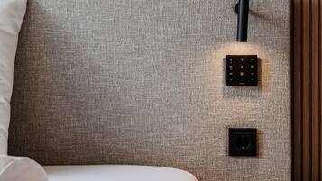 Una cama bien hecha con un cabecero gris, una mesita de noche con un control remoto y una lámpara de pared.