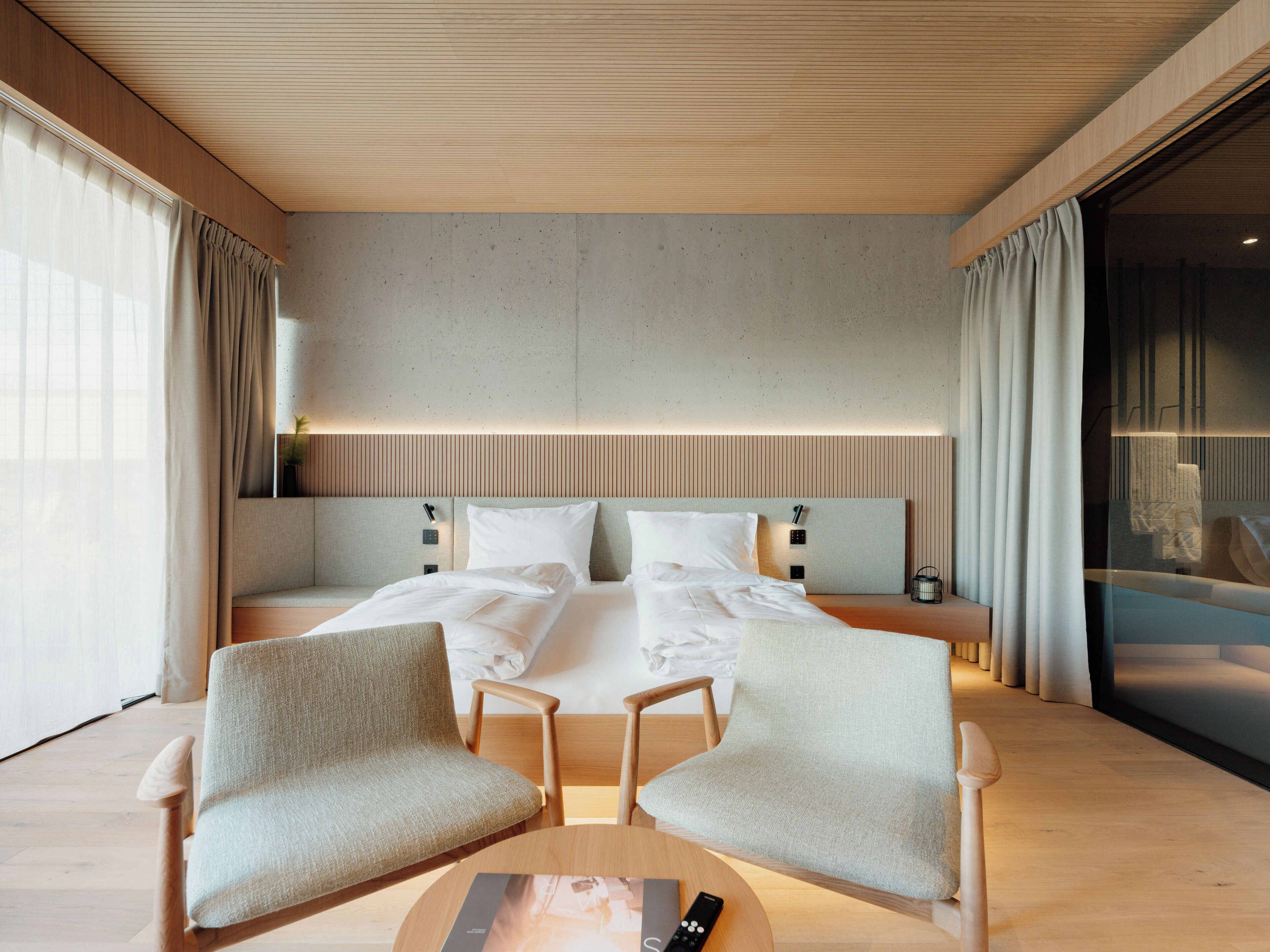Een moderne hotelkamer met een groot bed, twee fauteuils, een klein tafeltje en uitzicht op de stad.