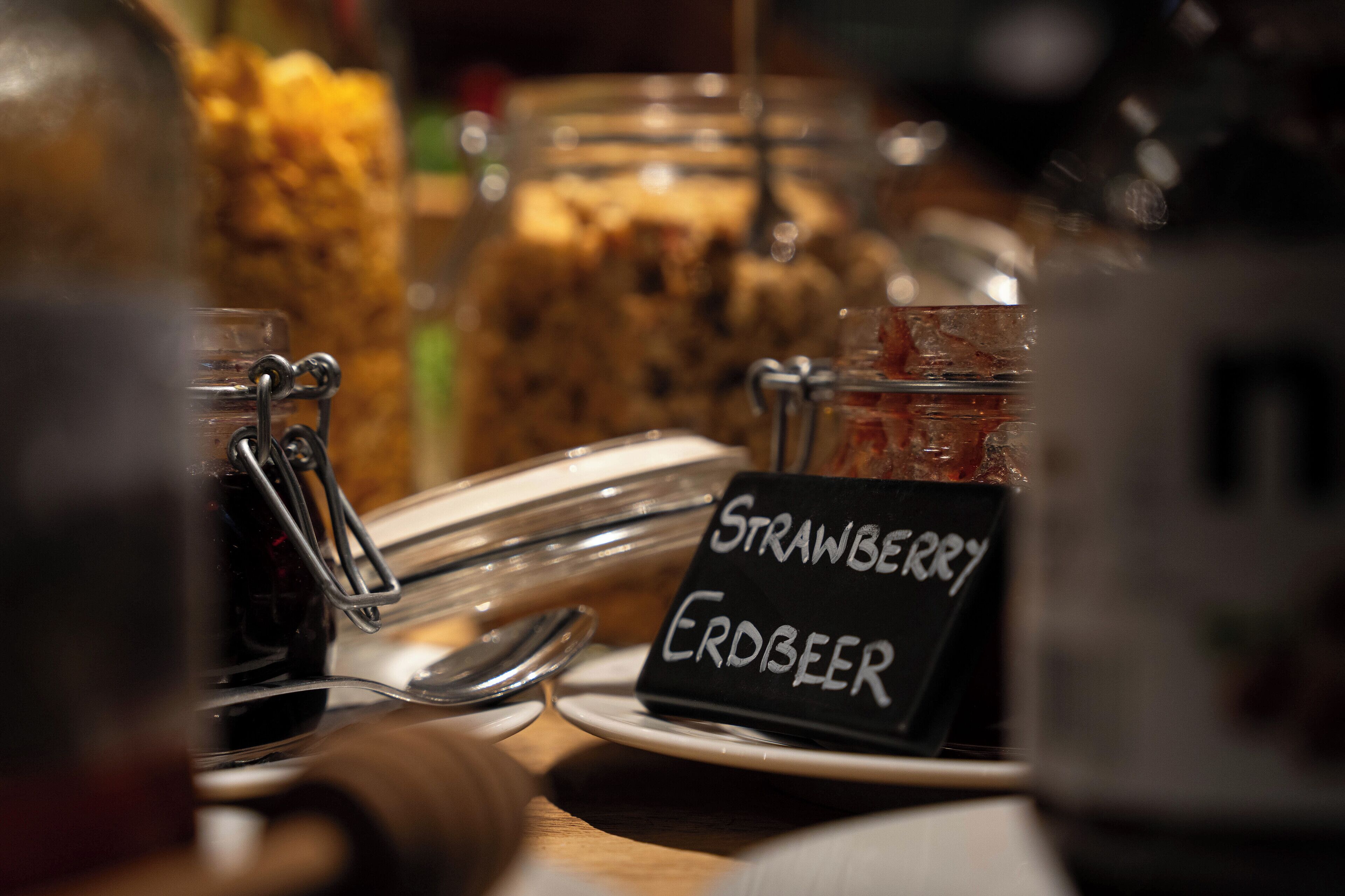 Ein Glas Erdbeermarmelade mit Etikett, ein Löffel und ein Glas im Hintergrund.