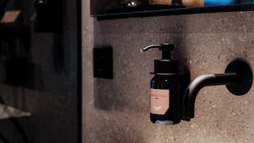 A bathroom with a black soap dispenser mounted on a wall, a small shelf above it, and a potted plant.