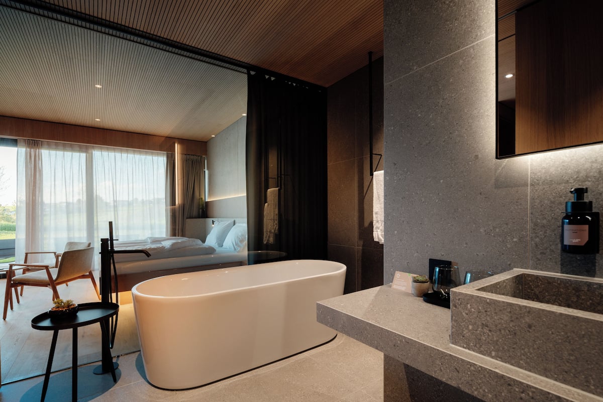 A modern hotel room with a large bathtub, a separate sitting area with a chair, and a large window offering a view.