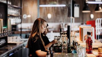 Zona de bar con un cantinero preparando bebidas, estantes con botellas y una barra con vasos y una caja registradora.