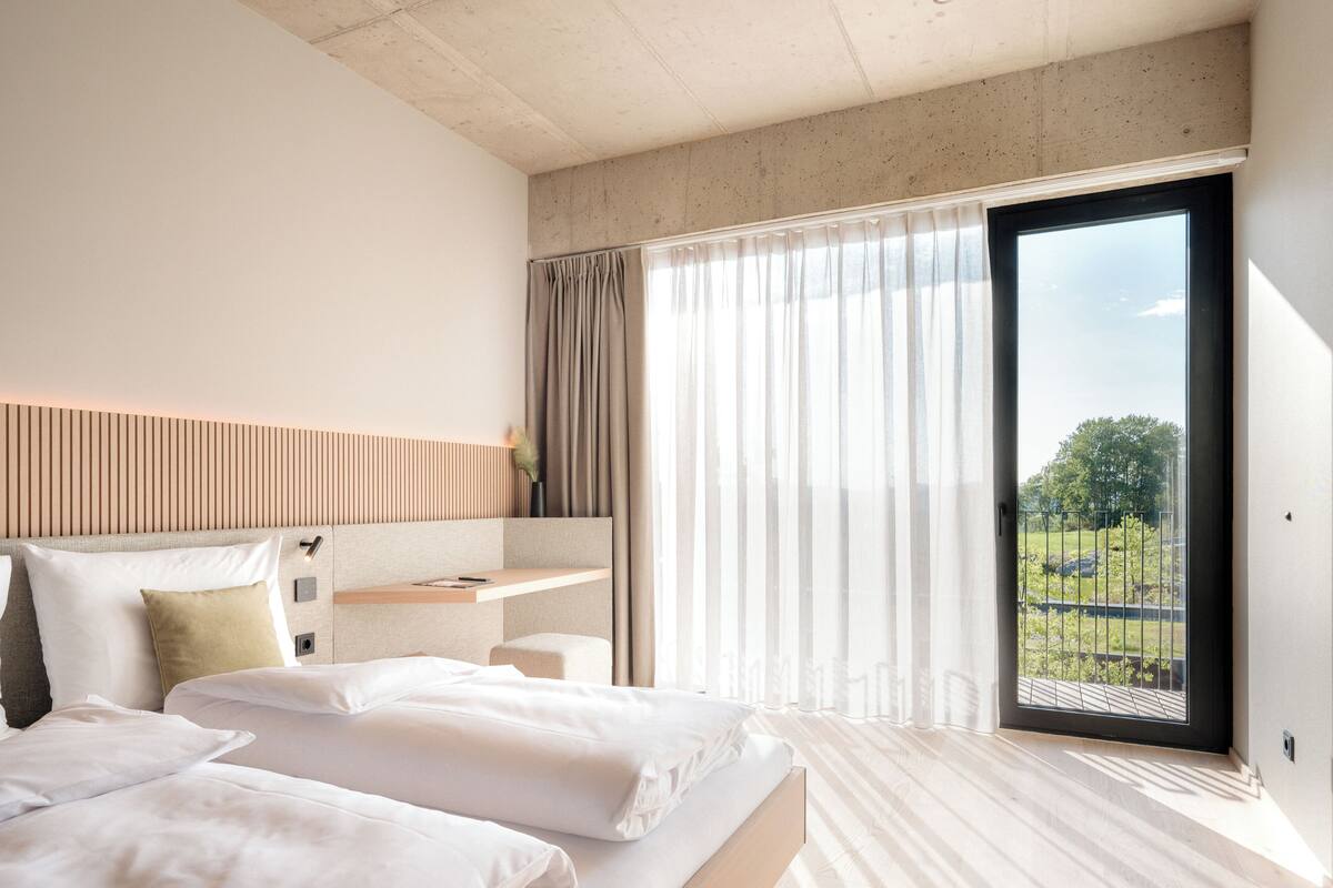 A modern hotel room with a large bed, a desk, and a view of a green landscape through a large window.
