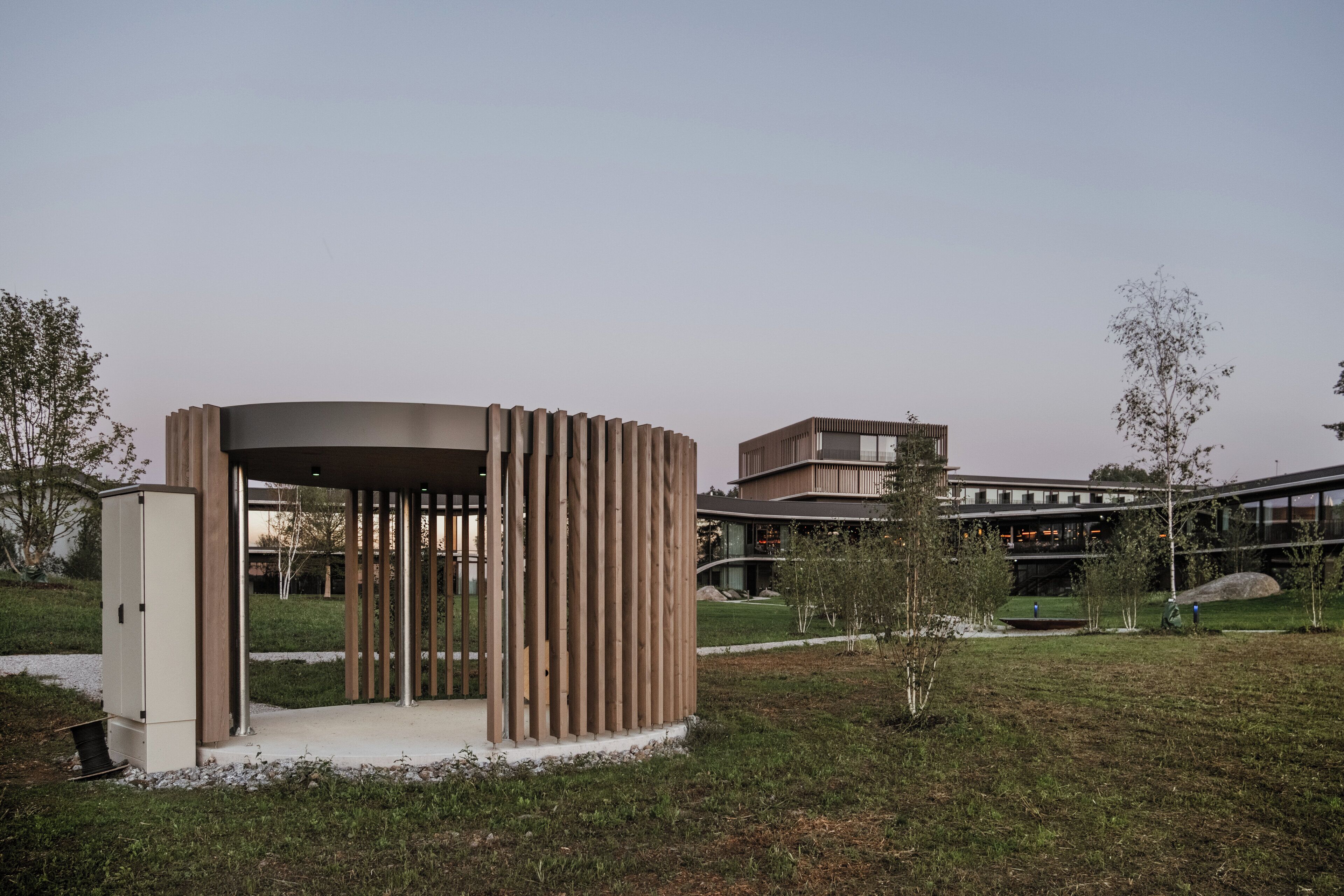 A modern pavilion with a curved roof and vertical wooden slats, surrounded by a grassy area and a building with a curved facade in the background.