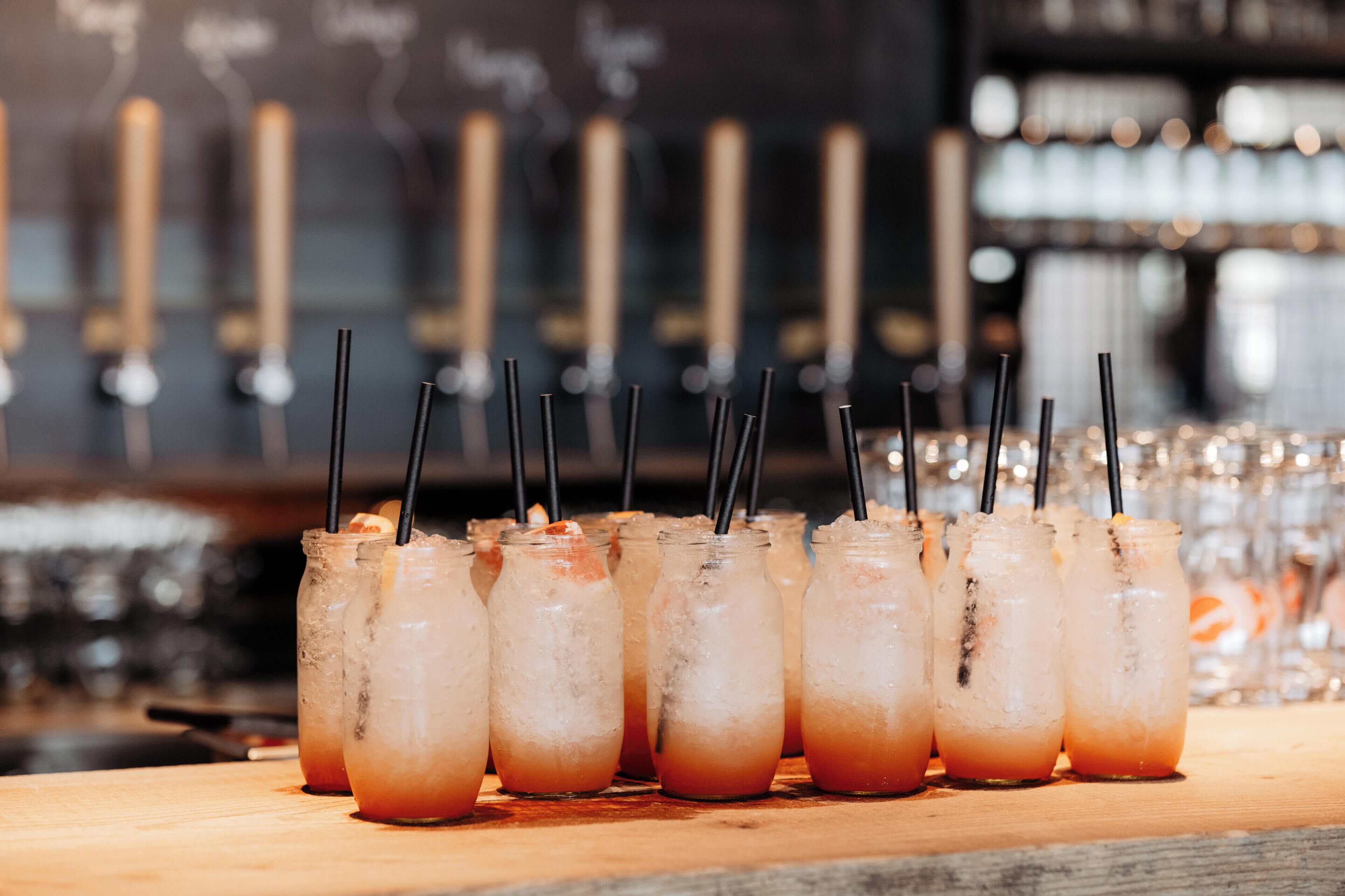 Un bar con varios vasos de bebidas heladas con pajitas, una barra de madera y un fondo borroso de estanterías.