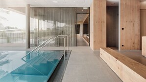 Un espace piscine intérieur moderne, doté de rampes en verre, d’un comptoir d’accueil en bois et offrant une vue sur l’extérieur grâce à de grandes fenêtres.