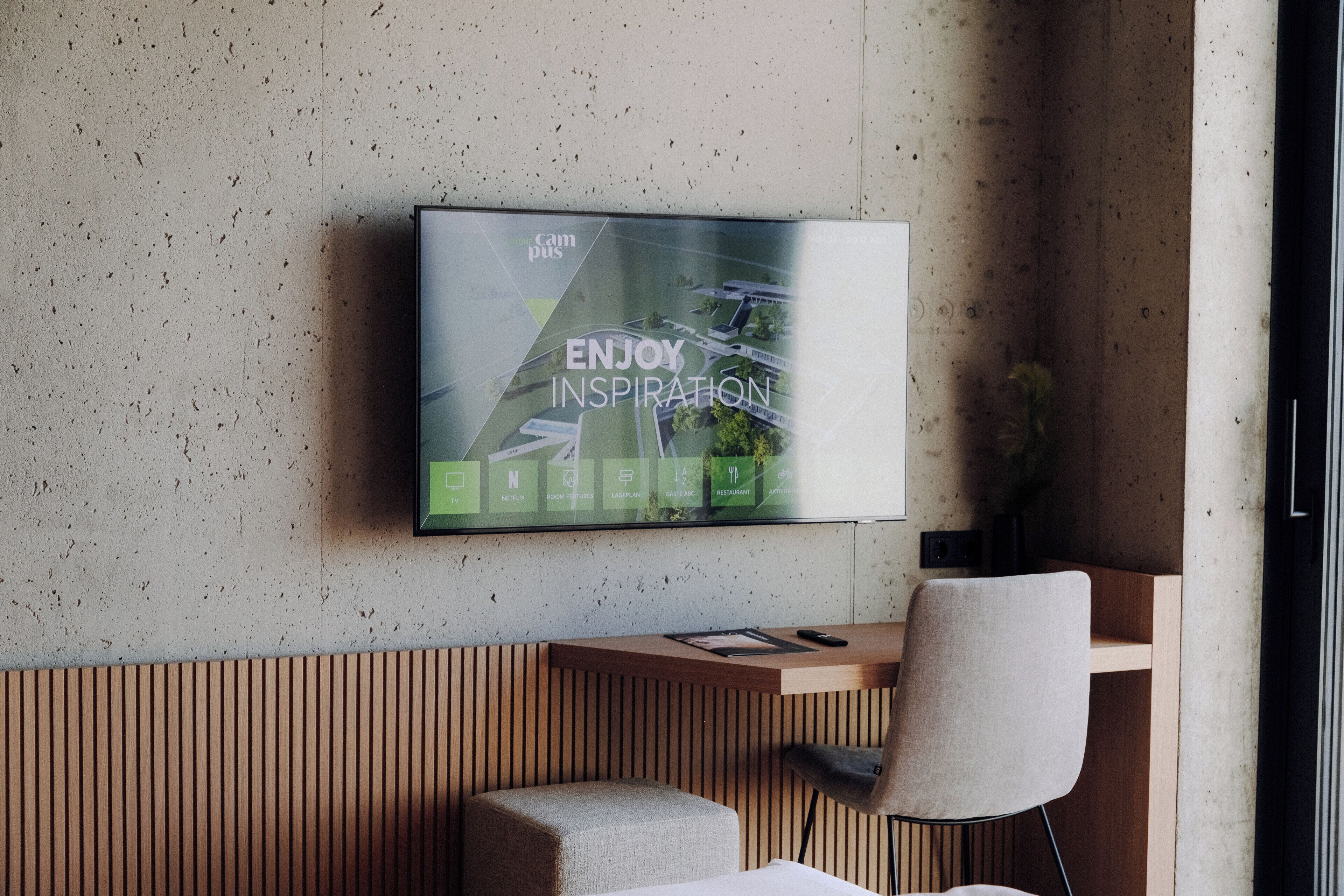 Et moderne hotelværelse med et fladskærms-tv monteret på væggen, et træskrivebord, en polstret bænk og en stol.