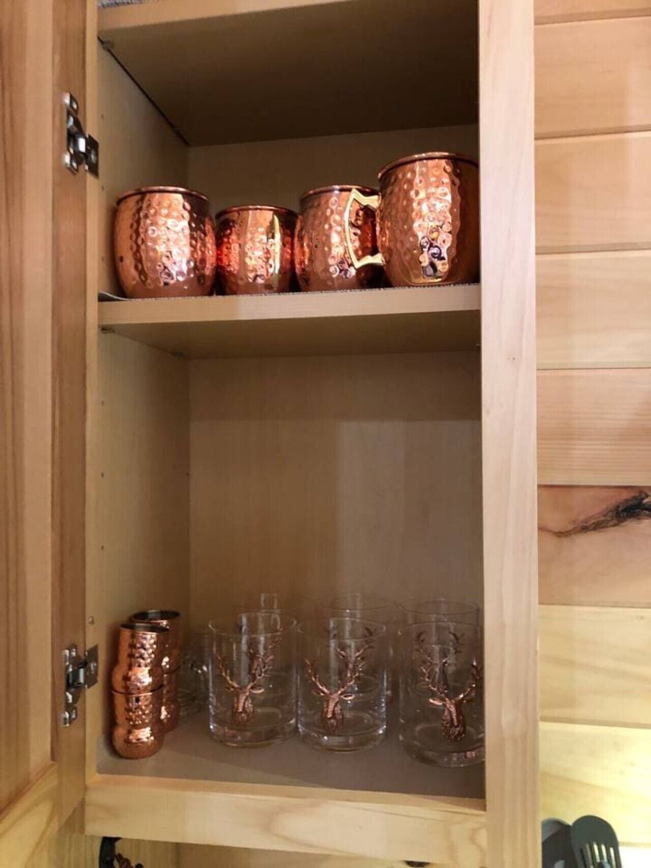 A wooden cabinet with two shelves; the top shelf holds copper mugs with a textured pattern, and the bottom shelf contains clear glass mugs with deer designs.