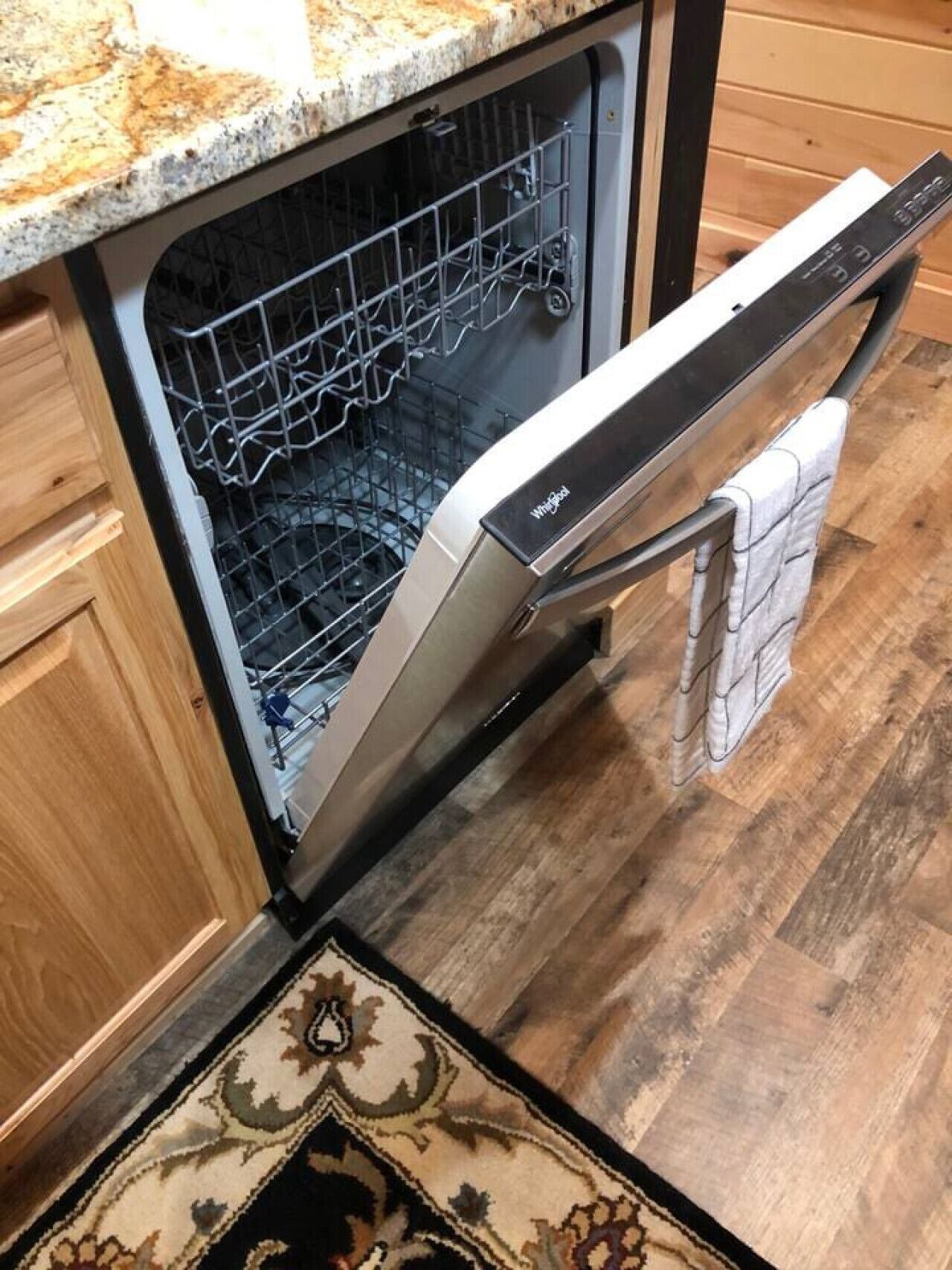 An open dishwasher in a kitchen with wooden cabinets and a patterned rug on the floor.