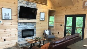 A log cabin interior with a stone fireplace, a flat-screen TV mounted above it, a leather sofa, and a dining table.
