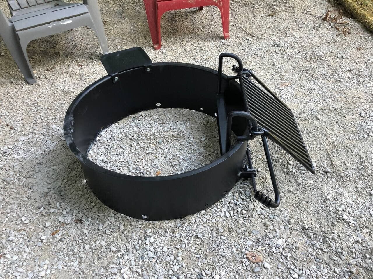 En rund grill til udendørs brug med metalrist, placeret på en grusbelagt overflade.