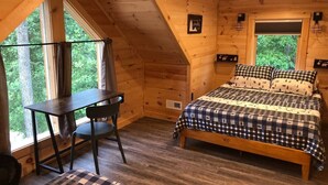 A cozy cabin room with a wooden ceiling, a bed with checkered bedding, a desk with a chair, and large windows offering a view of the forest.