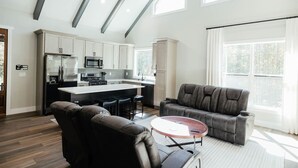 A modern kitchen with white cabinets, a central island, and stainless steel appliances. There is a leather sofa and a round coffee table in the living area. - Cedar Ridge Red River Gorge HotTub WiFi (Campton)