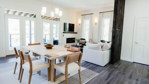 Een moderne woonkamer met een witte bank, een eettafel met stoelen, een aan de muur bevestigde televisie en grote ramen met gordijnen.