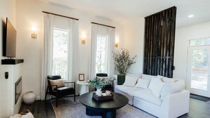 Een moderne woonkamer met een witte bank, een zwarte salontafel en een zwarte leren fauteuil. Er is een open haard, een aan de muur bevestigde televisie en een groot raam met gordijnen.