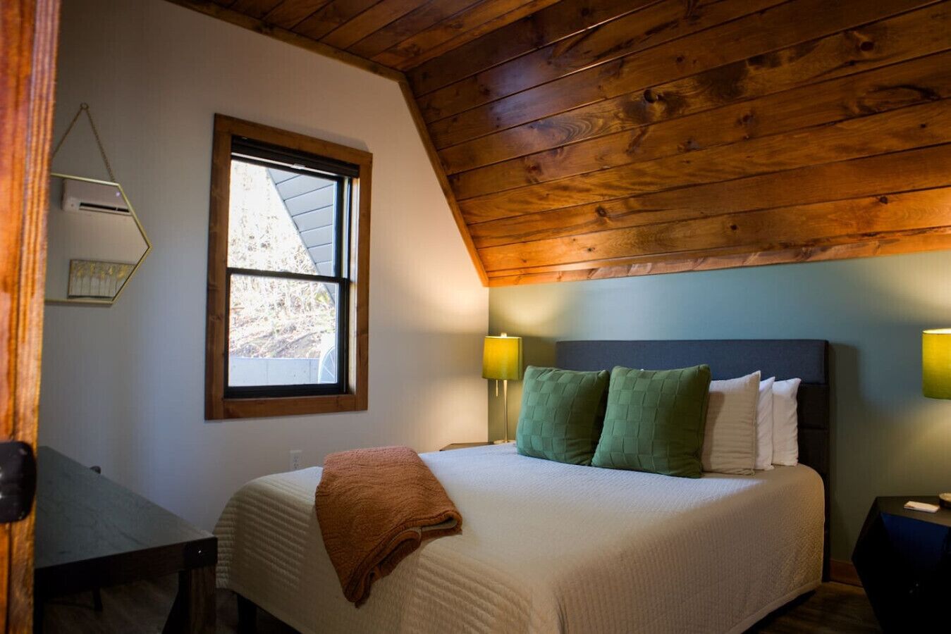 Een slaapkamer met een houten plafond, een bed met groene en witte kussens, een bruine plaid en twee bedlampjes.
