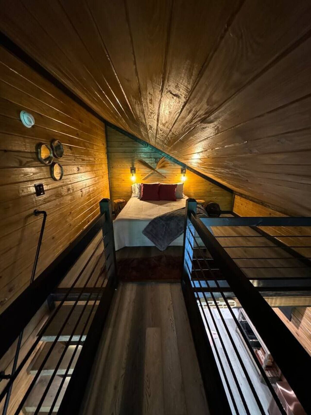 Un dormitorio bajo los techos, con techo de madera, una cama litera, dos luces fijadas en la pared y un ventanal pequeño.