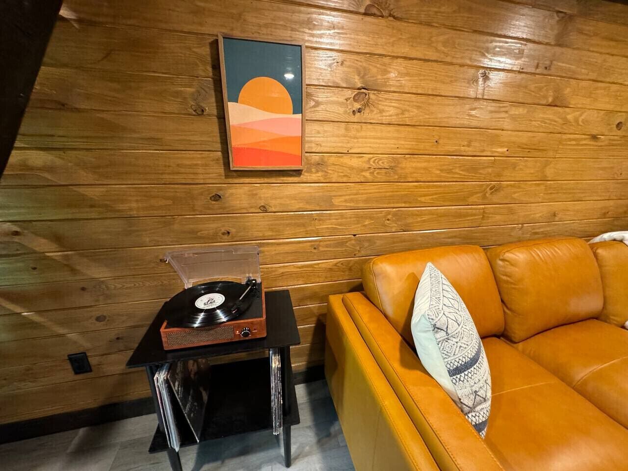 Et hyggeligt rum med en trævæg, en beige lædersofa, et lille sort bord med en pladespiller og et indrammet kunstværk over sofaen.