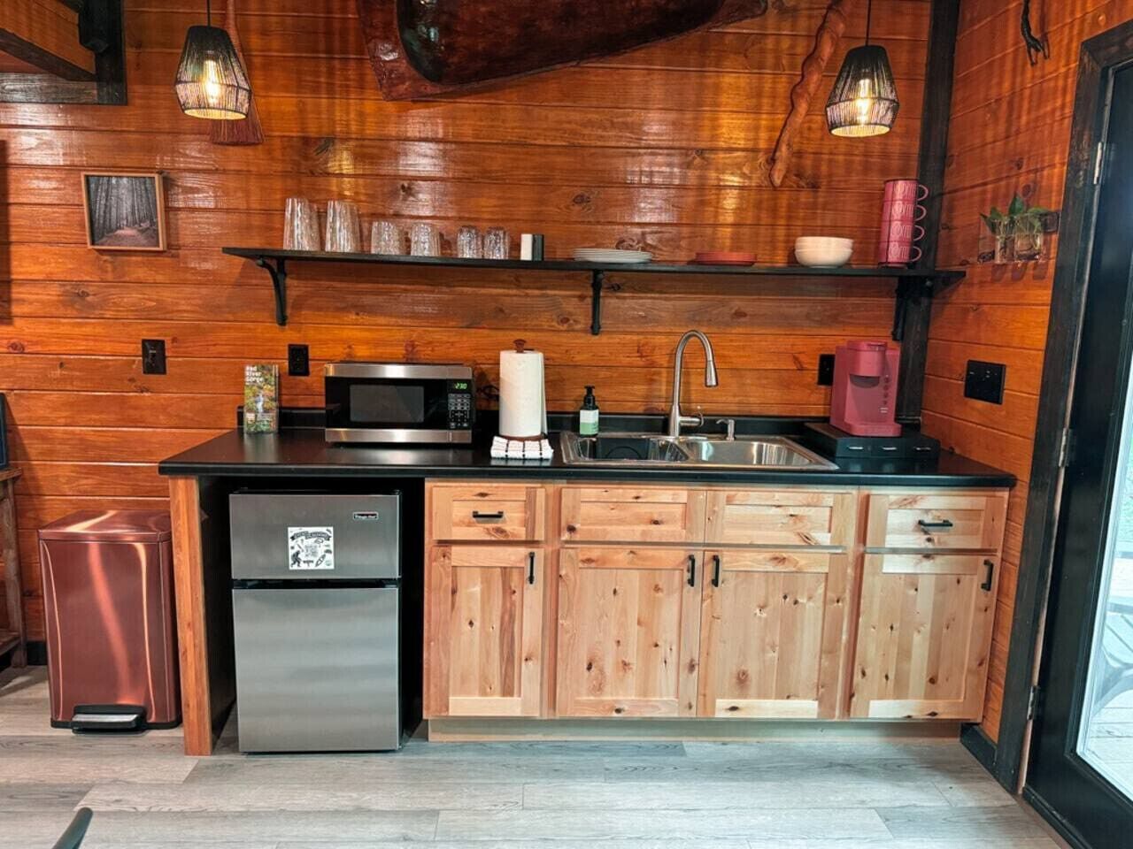 A compact kitchen with wooden cabinets, a stainless steel refrigerator, a microwave, a sink, and hanging pendant lights.